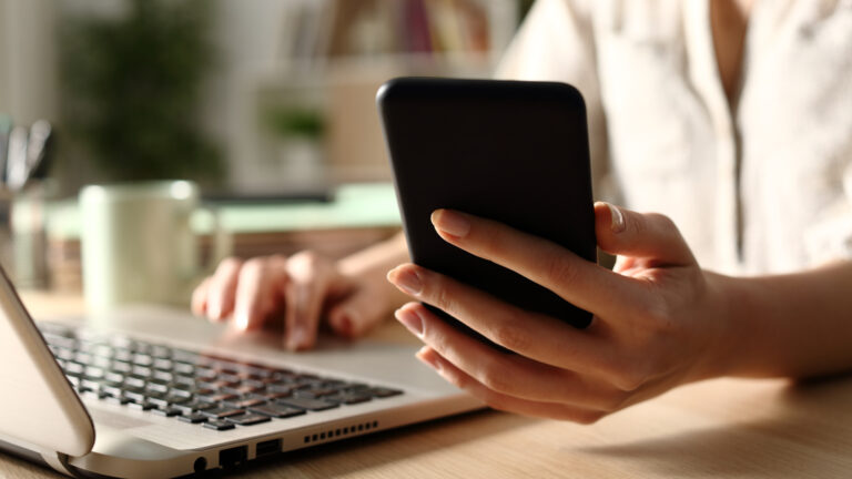 Close up of a cell phone held by a woman sitting in front of a laptop.