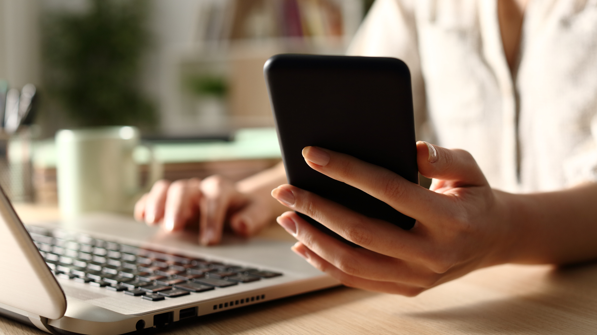 Close up of a cell phone held by a woman sitting in front of a laptop.