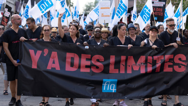 A crowd of people fill the street carrying a wide black banner with big red letters that say “Y’a des limites.” “There are limits.” The workers are all dressed in black shirts and many are waving small blue and white flags with the initials of their union, the FIQ.