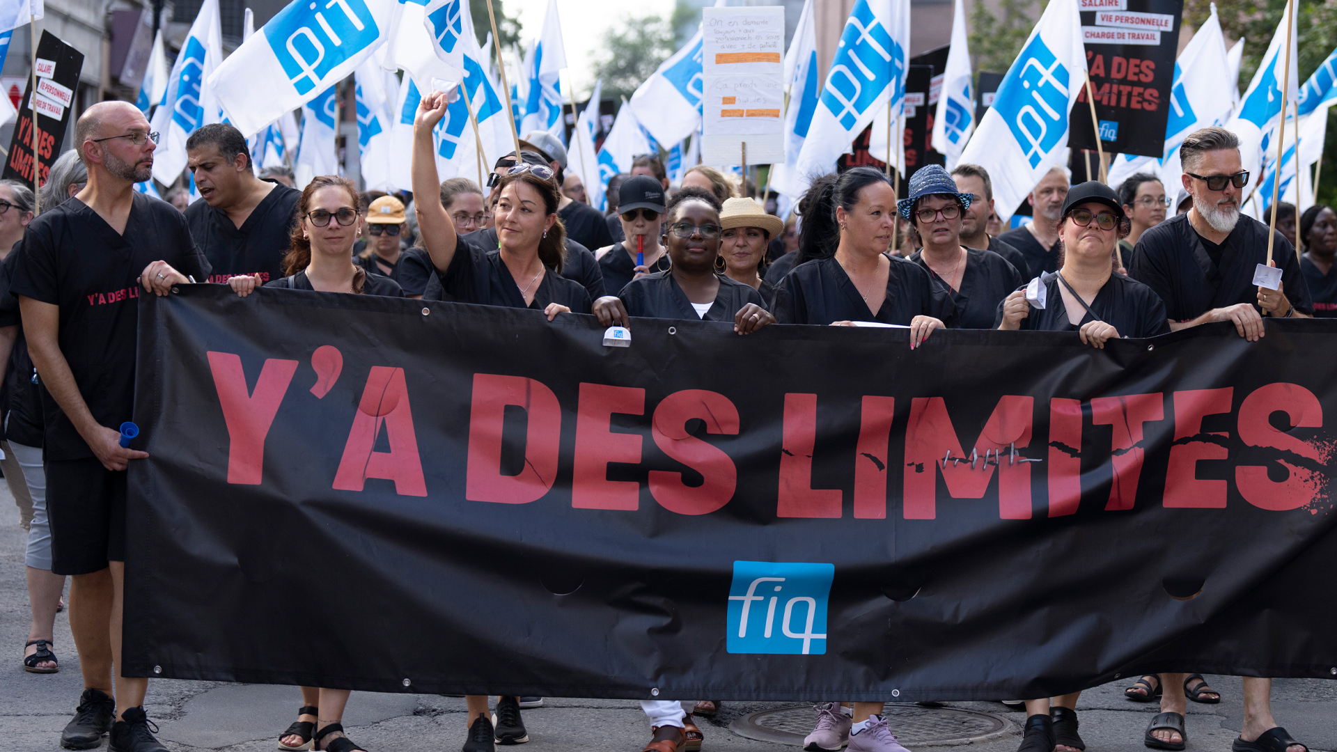 A crowd of people fill the street carrying a wide black banner with big red letters that say “Y’a des limites.” “There are limits.” The workers are all dressed in black shirts and many are waving small blue and white flags with the initials of their union, the FIQ.