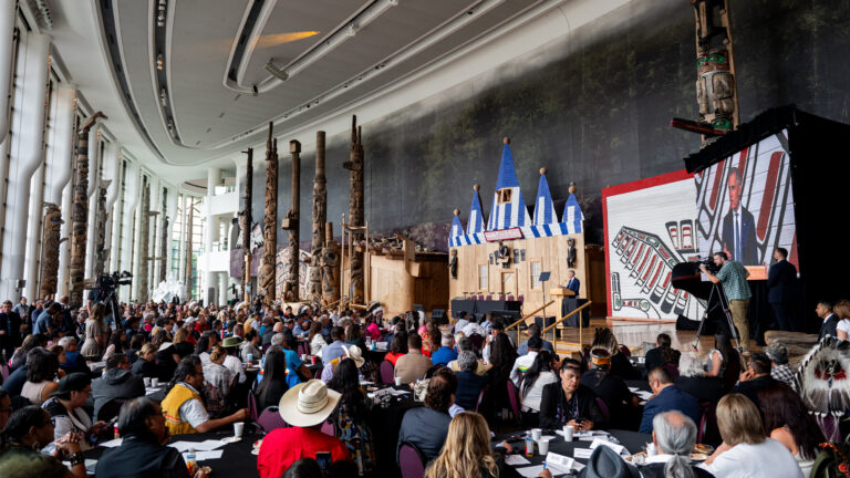 Mark Carney in the distance speaking to a crowd of people in a room decorated with Indigenous symbols such as paintings and totem poles.