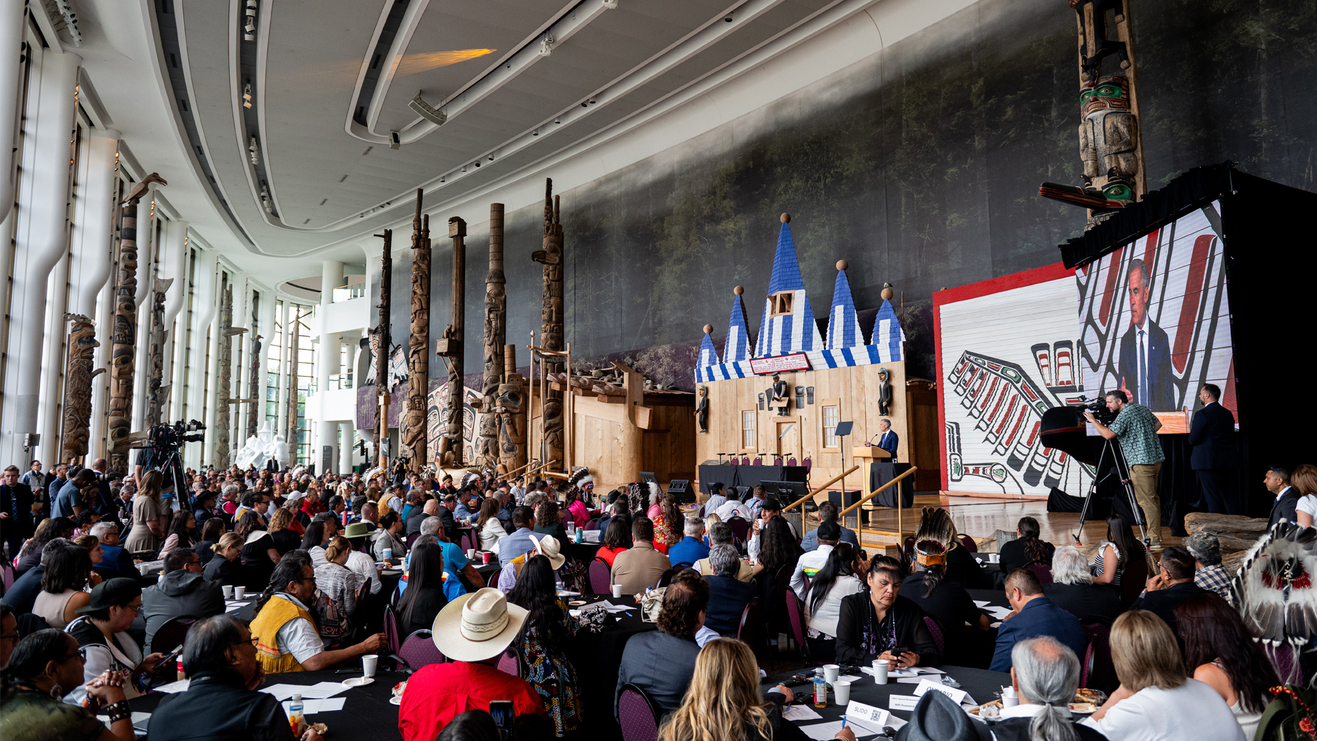 Mark Carney in the distance speaking to a crowd of people in a room decorated with Indigenous symbols such as paintings and totem poles.