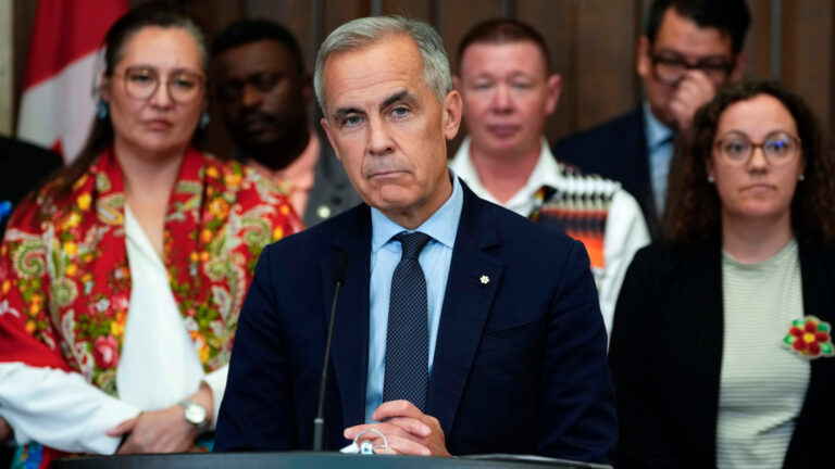Carney stands before a podium with his hands clasped in front of him. Five cabinet ministers are slightly out of focus in the background.