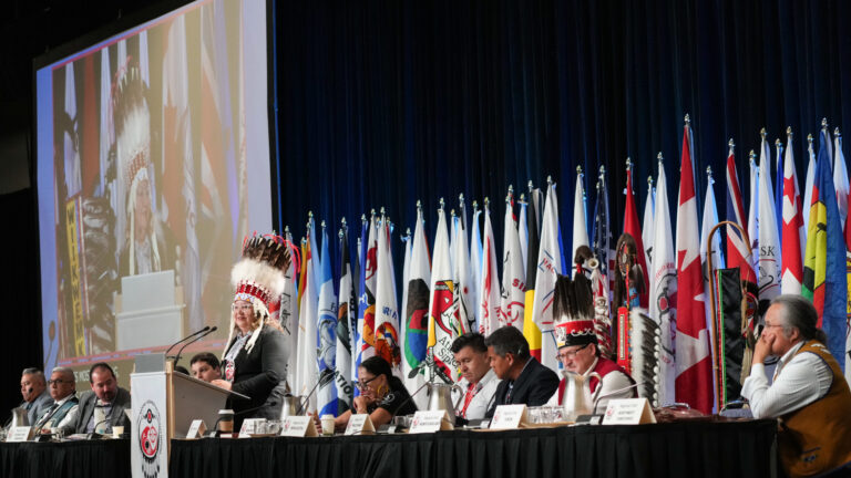 Indigenous National Chief Cindy Woodhouse speaking at podium during an assembly, surrounded by First Nations representatives and flags.