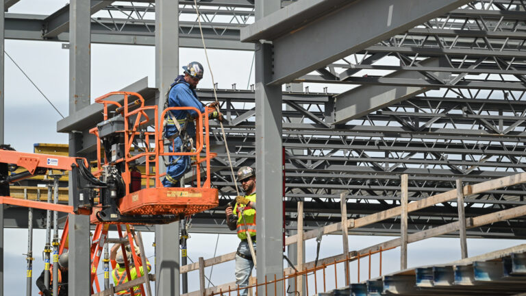 A man in a hard hat is adjusting a wire being held by a co-worker. The man is standing on an orange metal platform near the grey metal beams and scaffolding of the building.