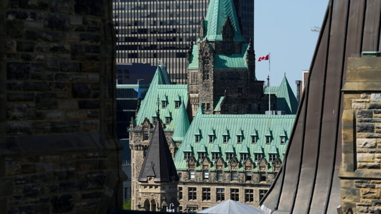 The peaked pistachio-green rooftops of the stately old stone government buildings sit in contrast to the taller towers around them. A Canadian flag flies on a mast from the roof of the Confederation Building.