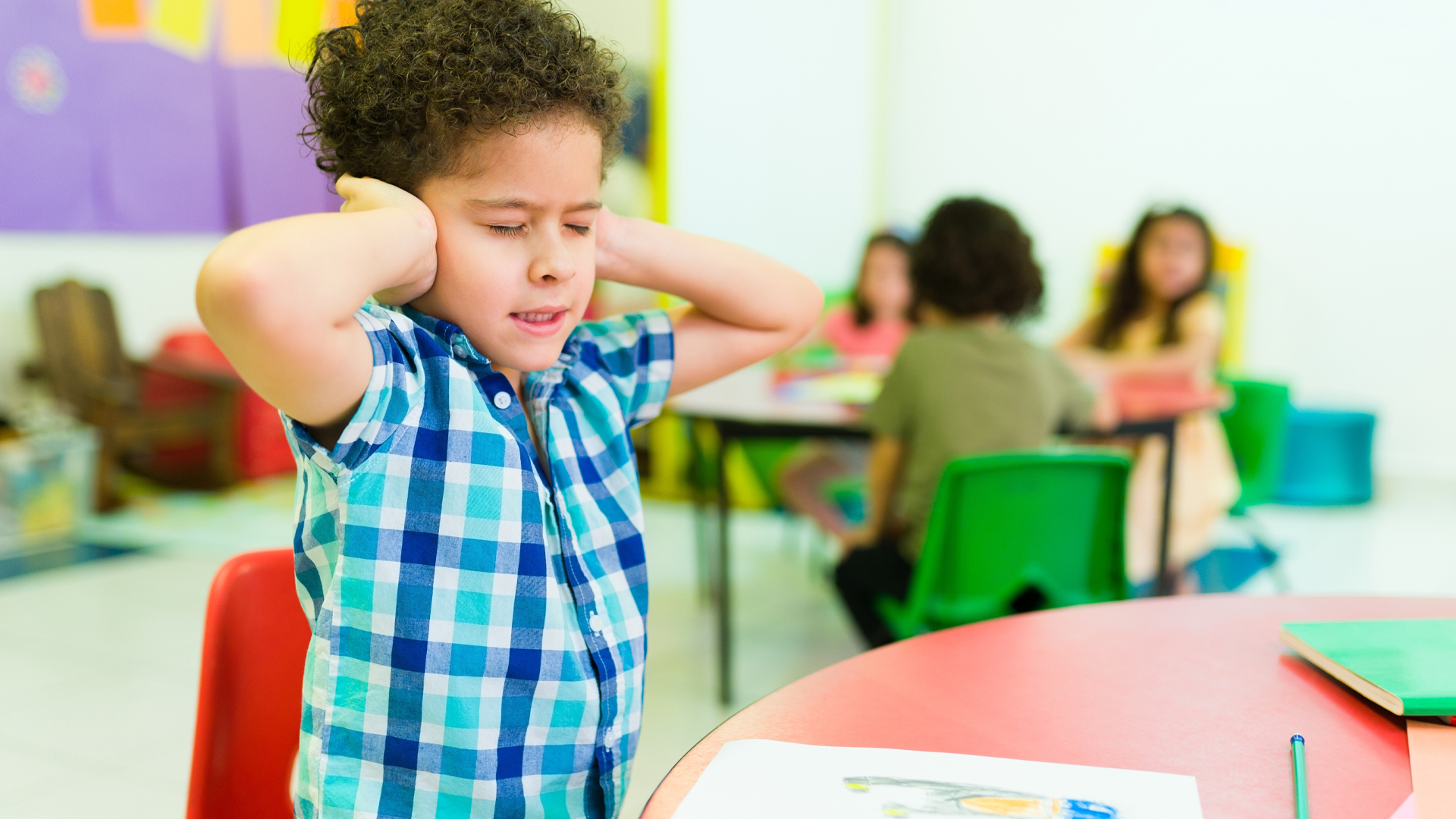 A primary school aged boy covers his ears in a noisy classroom.