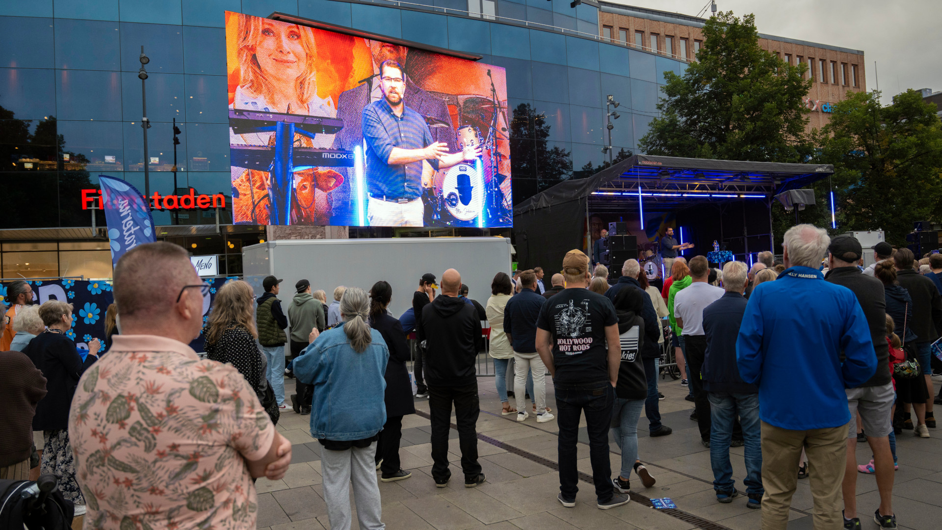 People gathered outside in the public square watch a giant screen on which is projected the image of a man in a blue shirt talking.