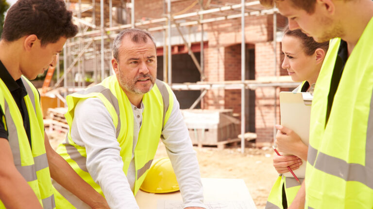 Middle-aged white man points to paper plans as three younger white workers (two men, one woman) look on. They are all wearing fluorescent green safety vests, standing near a brick building with scaffolding.