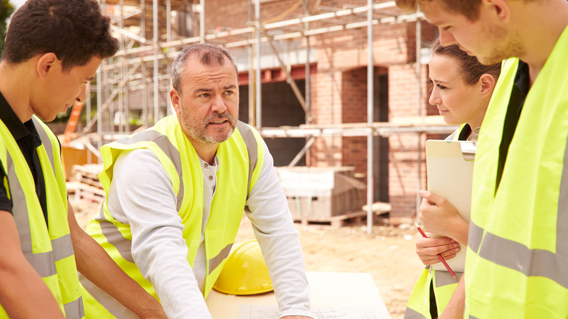 Middle-aged white man points to paper plans as three younger white workers (two men, one woman) look on. They are all wearing fluorescent green safety vests, standing near a brick building with scaffolding.