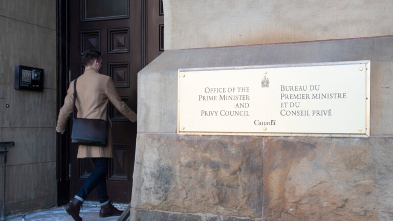 A person in a beige coat walks through a front door to the left. To the right is a bronze plaque that says “Office of the Prime Minister and Privy Council.”