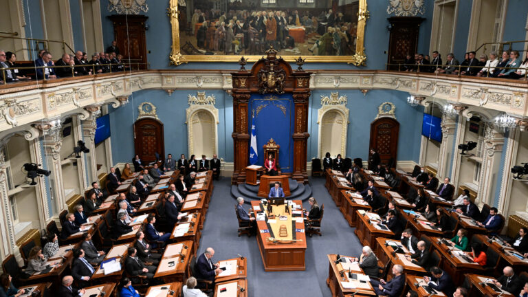 People sit at rows of desks oriented toward the middle of the room, where a central desk and the Quebec flag are located. A second-floor gallery is populated with a row of observers. At the back of the room is a large gilt-framed painting, in keeping with the ornate details that make up the room.