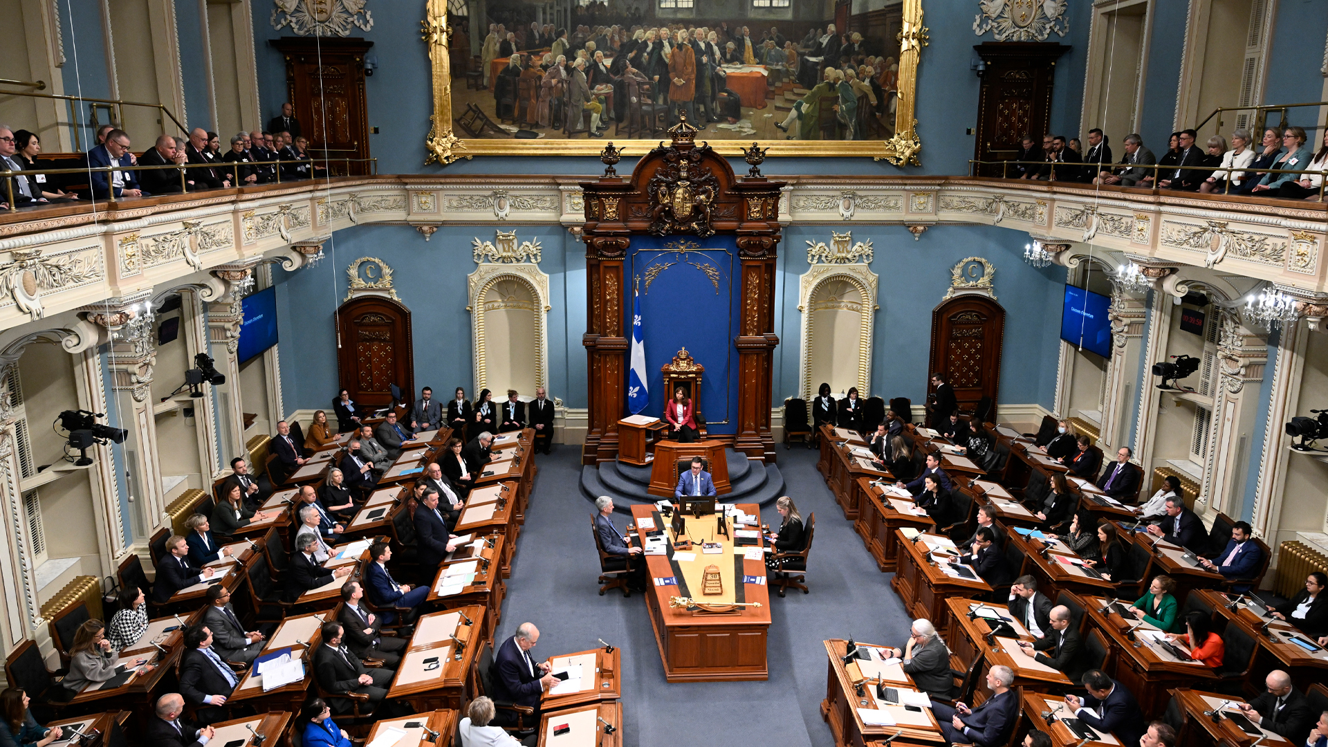 People sit at rows of desks oriented toward the middle of the room, where a central desk and the Quebec flag are located. A second-floor gallery is populated with a row of observers. At the back of the room is a large gilt-framed painting, in keeping with the ornate details that make up the room.