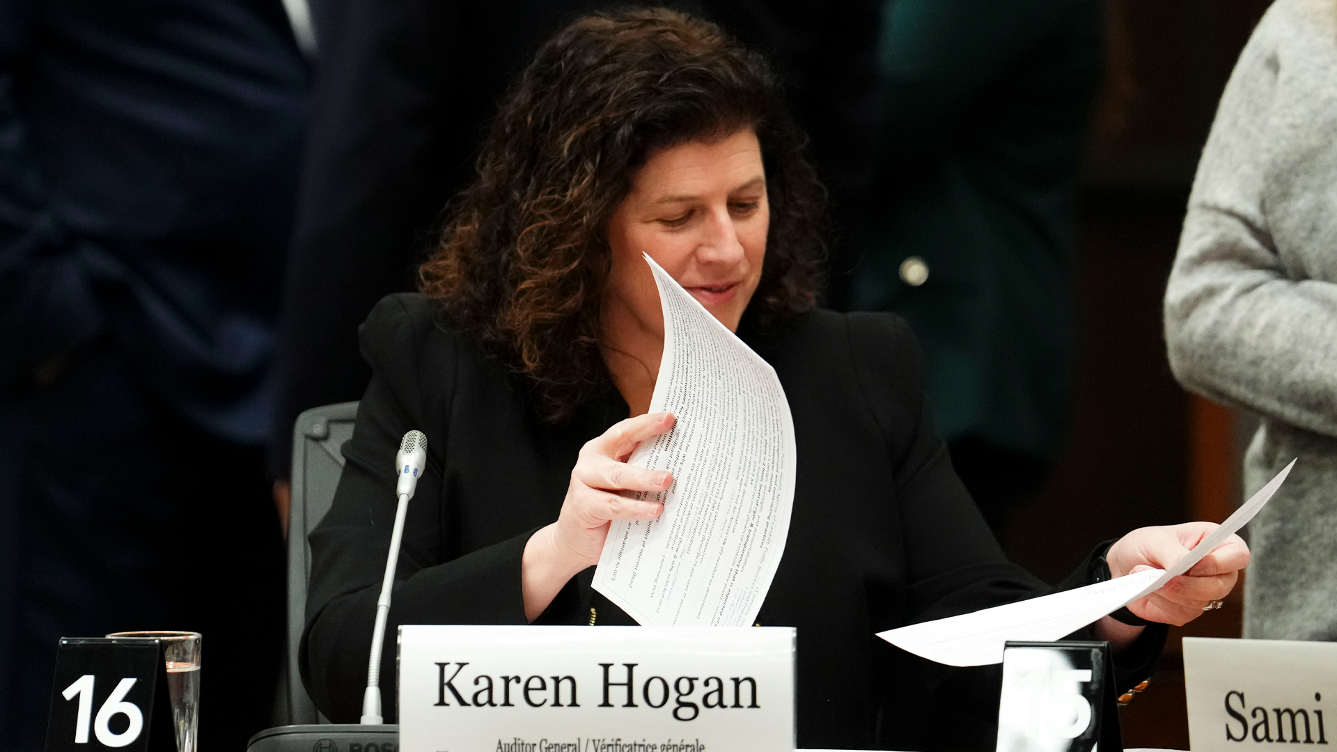 Hogan is seated at a long wood table with a paper nameplate and a slender silver microphone in front of her. She is looking at papers, and is holding on to two single sheets as she looks at the pile.