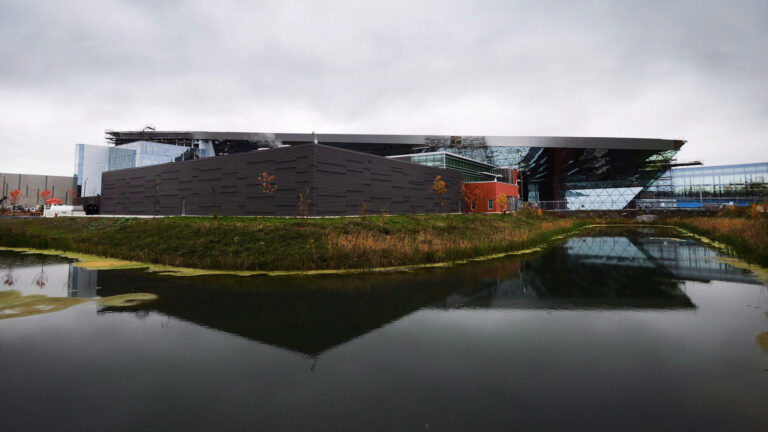 A sleek modern building with dark brown brick in the foreground and glass in the background viewed from across a small pond.