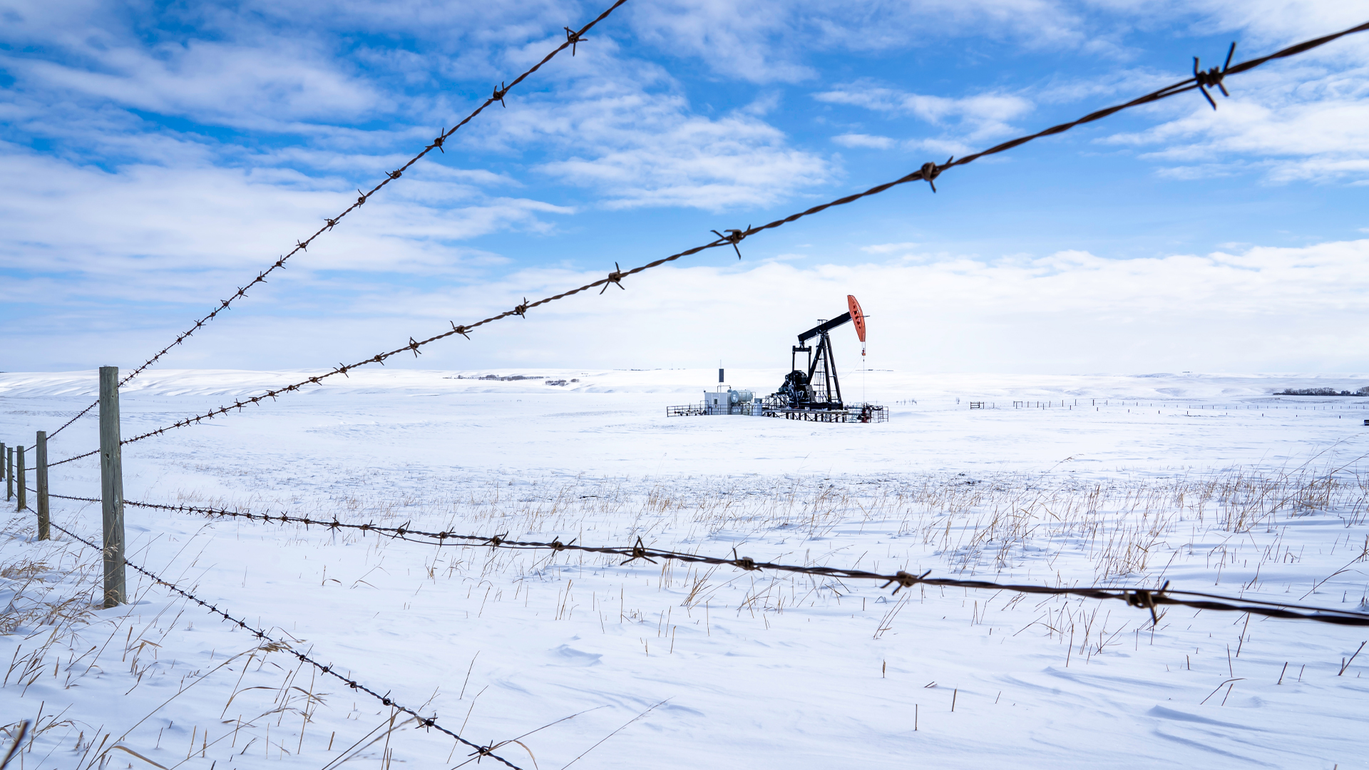 Un puits de pétrole dans un champ couvert de neige, vu à travers une rangée de barbelés.