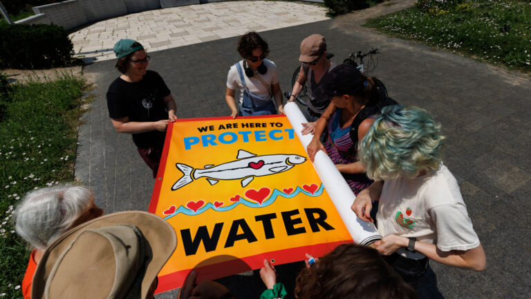 Eight people hold the edges of a banner that says “We are here to protect water.” The banner is orange, blue, red and white, and has an illustration of a fish with a heart.