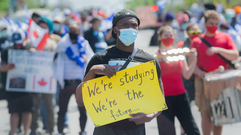 A man holds a yellow sign saying “We’re studying, we’re the future…” with other protesters in the background.