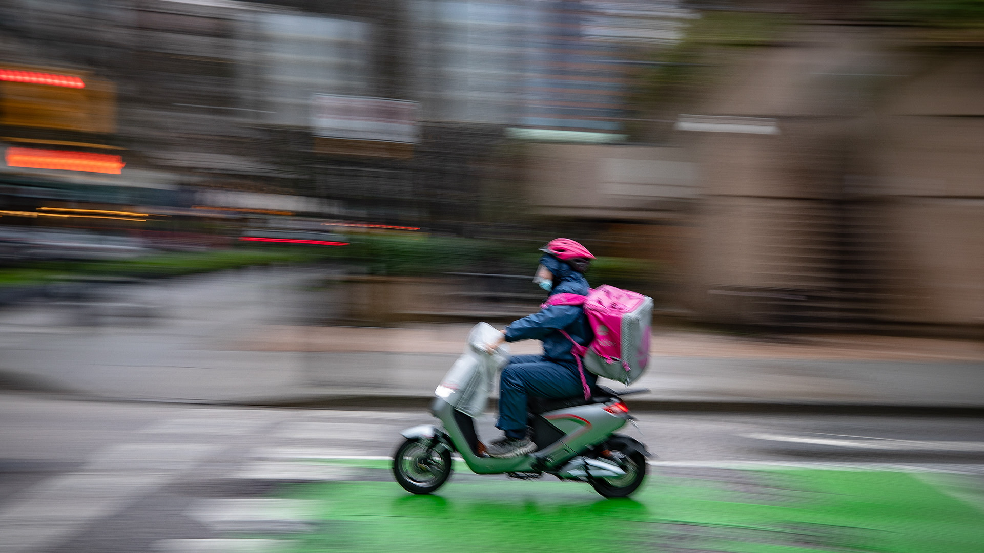 Un livreur de nourriture avec un casque rose et un sac à dos isolant carré rose conduit un scooter à Vancouver.