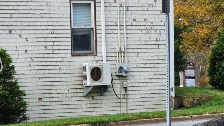 The unit is white and tan and is mounted on a bracket sitting several feet off the ground. The building has light grey wooden siding, and a single narrow window sits above the unit.