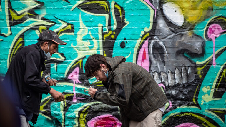 Behind the men is brick wall covered in a bright, turquoise-coloured mural depicting the partial skull of a creature with a large white eye and teeth. One man is hunched over and examining something in his hand. The other is watching what the man is doing.