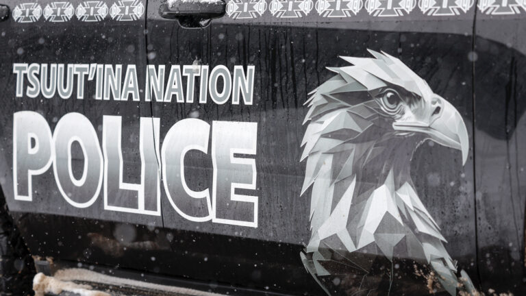 Une tête d’aigle stylisée orne la portière d’une voiture de police, en gros plan, à côté du nom de la Première Nation et du mot « police » écrit en très grosses lettres, en gris pâle sur fond noir.