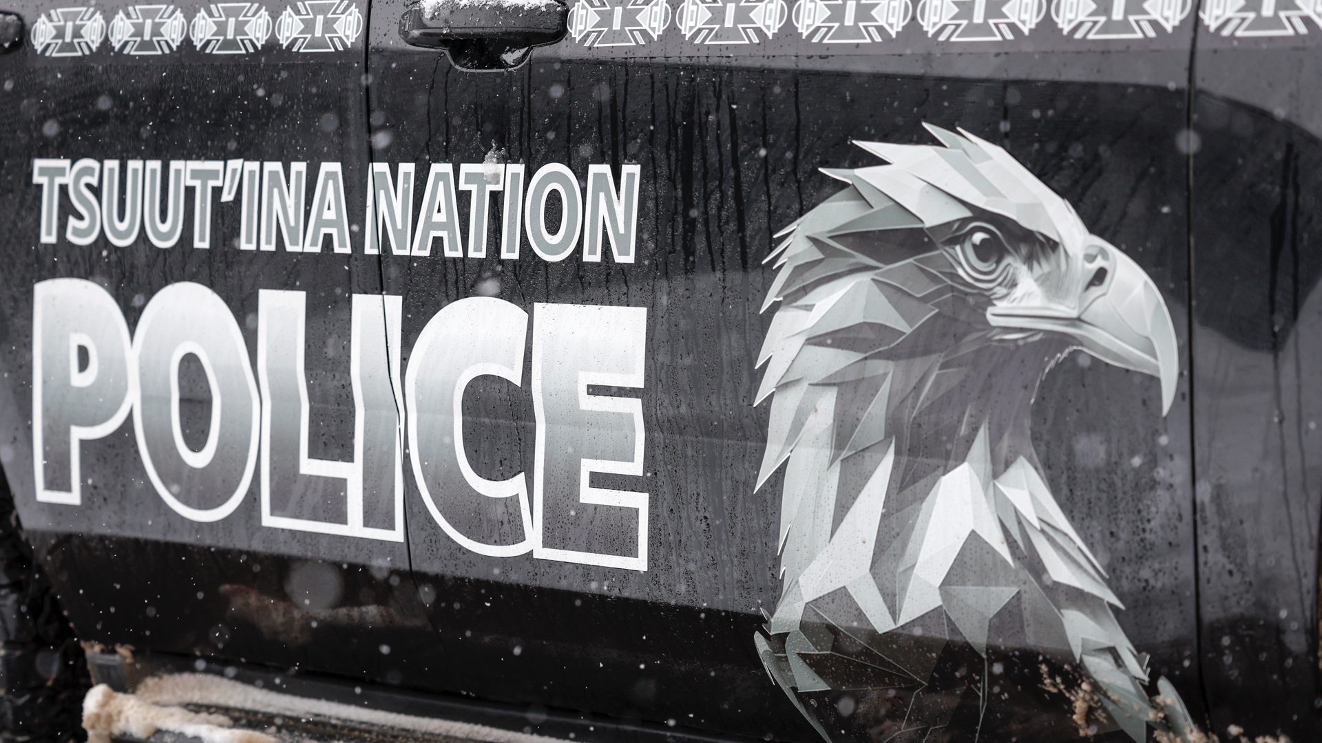 Une tête d’aigle stylisée orne la portière d’une voiture de police, en gros plan, à côté du nom de la Première Nation et du mot « police » écrit en très grosses lettres, en gris pâle sur fond noir.