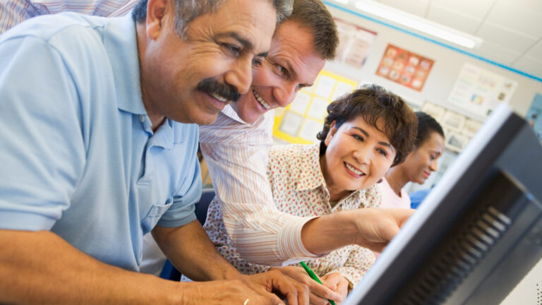 A row of four adults sits in front of computers. Two men and a woman smile and lean in to look at something on one screen. They are all at least 40. One man has greying hair. They are in a classroom with a wall covered in posters.