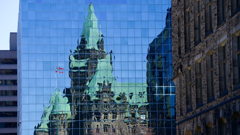 The heritage Confederation building is blurred in the reflection of a modern glass tower. 