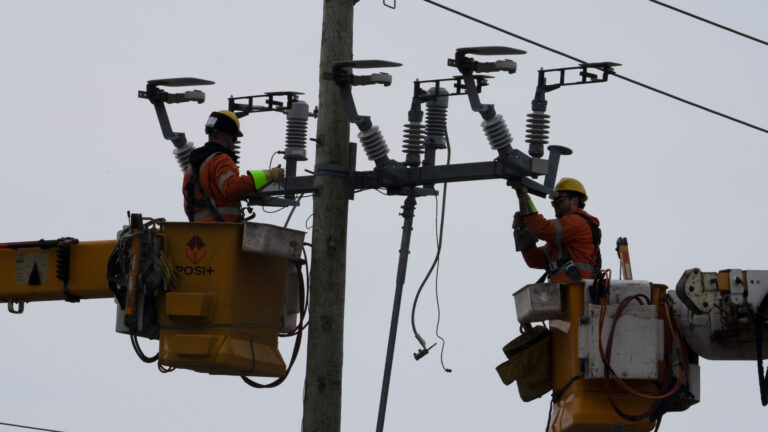 High in the air on a cloudy day, two men in orange uniforms and hardhats work on the fixtures at the top of a power pole connected to high-tension wires. Each man is standing in a mechanical bucket that’s been raised from a hydro truck. A few stray wires are dangling.