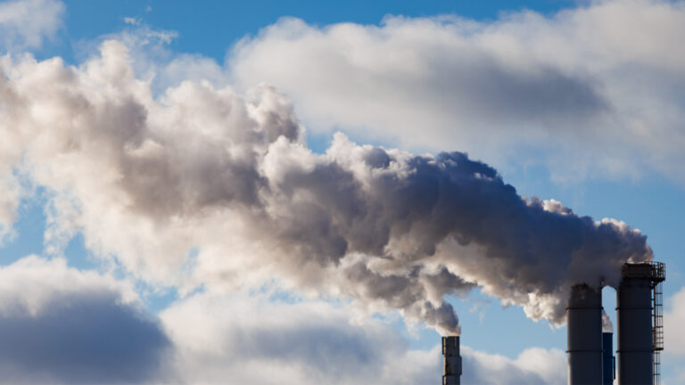 Smoke billows from three smokestacks against a blue sky with billowy clouds.