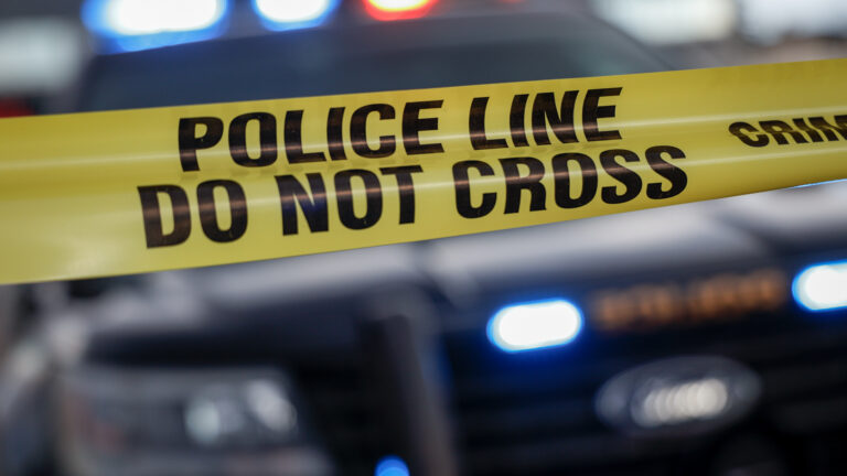 A yellow police tape marks the edge of a crime scene with a blurry black police car, red and blue cruiser lights on, in the distance. The tape says “Police line. Do not cross” in thick black letters.