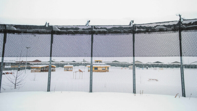 A snow covered jail yard is pictured behind a tall chain-link fence. The yard has several small shelters in front of a large single-storey complex.
