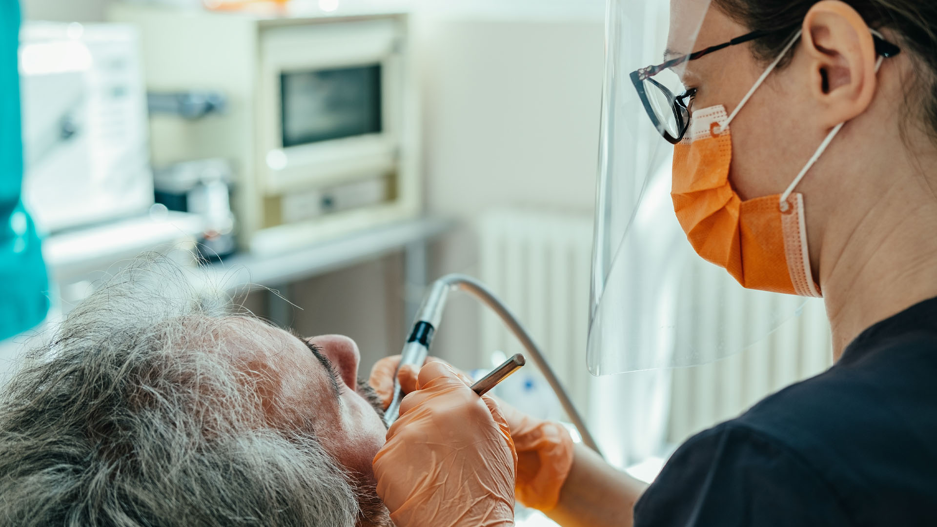 Un homme aux cheveux grisonnants reçoit des soins d'une professionnelle des soins dentaires portant des gants et un masque orange et une blouse bleu foncé.