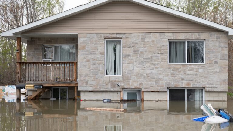 A beige, stone bungalow is surrounded by brown-ish water that is about five feet high. The basement-level doors and windows are three-quarters under water. Mature trees surround the house.