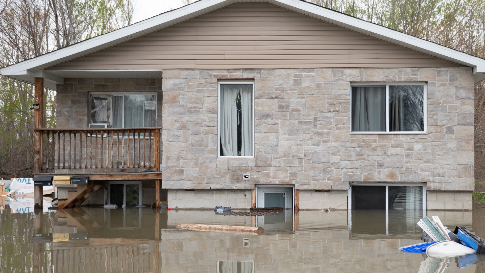A beige, stone bungalow is surrounded by brown-ish water that is about five feet high. The basement-level doors and windows are three-quarters under water. Mature trees surround the house.