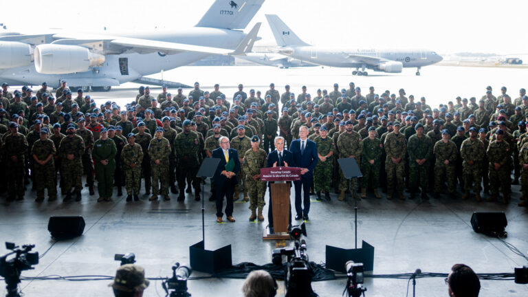 Carney stands at a podium on the tarmac. Behind him is several hundred soldiers in olive-green uniforms, and behind them are two white jets. A row of media and cameras on tripods is facing Carney.