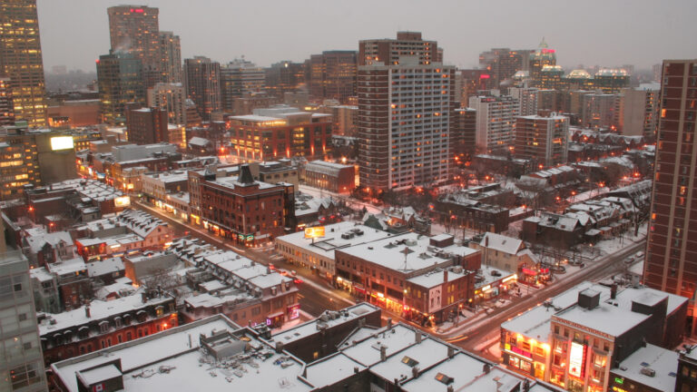View from a high-rise showing snow-covered roofs of homes and shops on a few blocks of downtown streets, with street lights glowing with the backdrop of a flat grey sky at dusk.