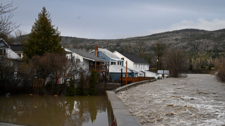 A river rushes past a temporary flood barrier attempting to protect several river-front houses. Standing water visible behind the barrier.