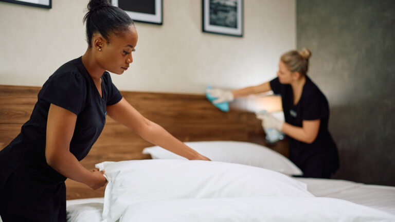 Two women in black uniforms make a bed in a hotel room. 