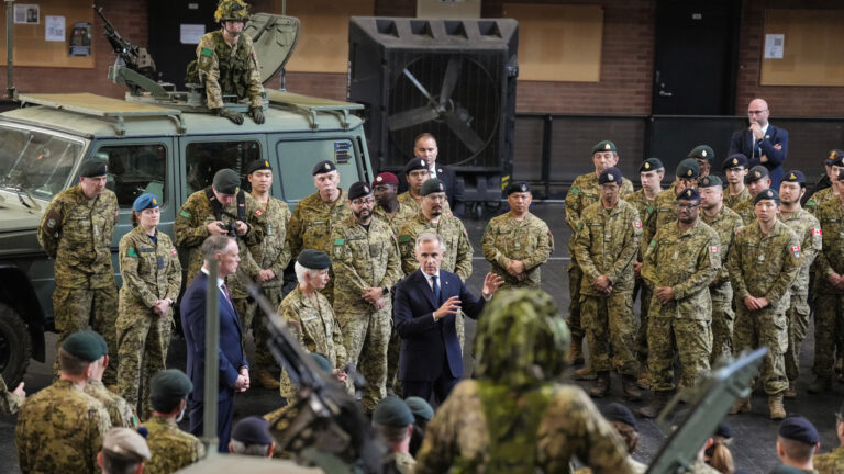Mark Carney, wearing a suit, is surrounded by soldiers in green camouflage fatigues and military vehicles.