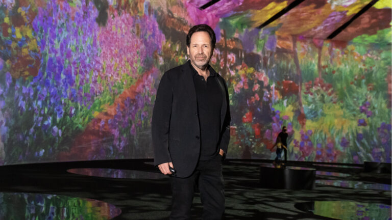 A portrait of a man dressed in a black coat and jeans in front of a multi-coloured backdrop.