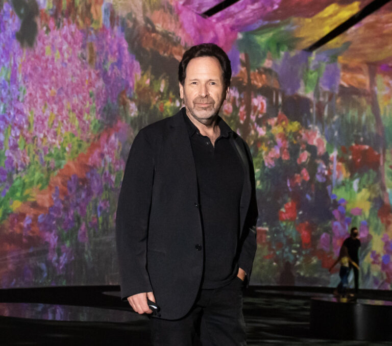 A portrait of a man dressed in a black coat and jeans in front of a multi-coloured backdrop.
