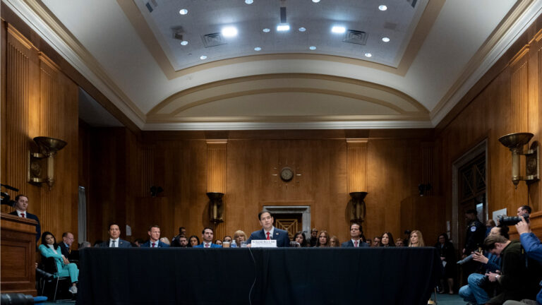 A wide shot of Marco Rubio sitting alone at a long table. With onlookers behind him.