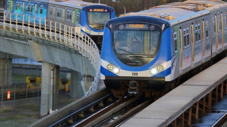 Two trains ride on outdoor above-ground rail lines, one of them on a curve on a track that runs below the other train. The fronts of the trains are blue with a white stripe at the bottom. Several passengers can be seen in the front window of one train.