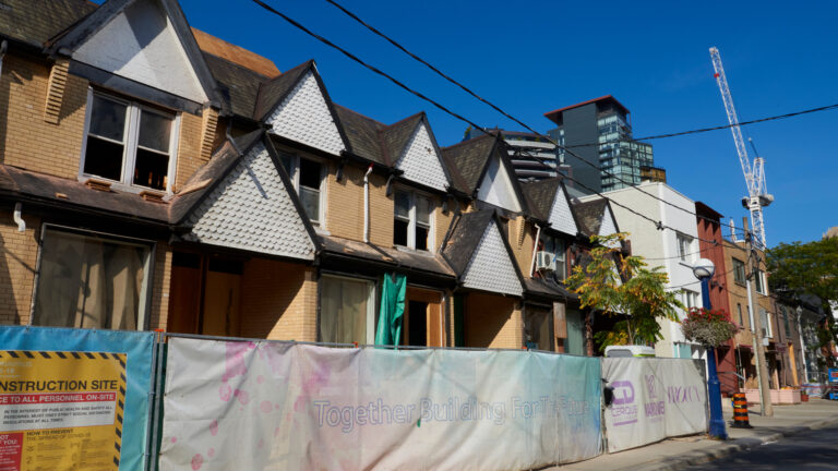 The rowhouses are two storeys high, with a pattern of gables interspersed between windows. Some of the window panes have been removed. Farther down the street are mature trees. In the distance are a couple of new towers.