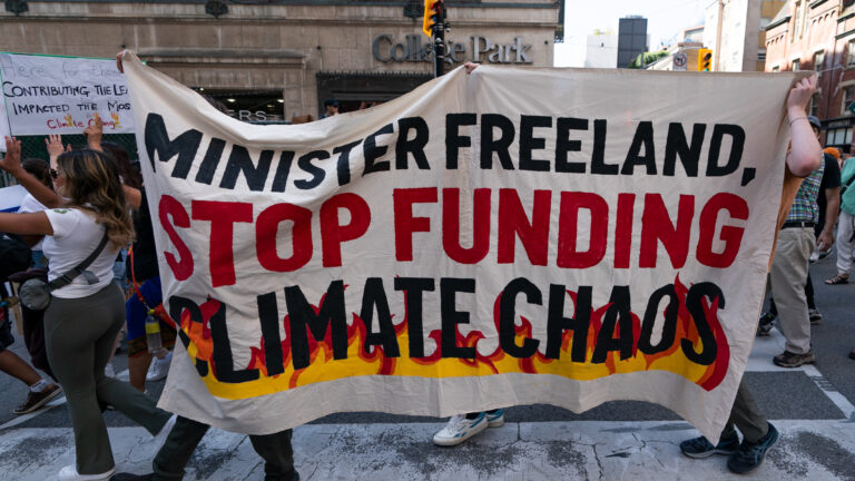 A crowd fills the street with some people holding signs. One large rectangular sheet of canvas is held up by three people. It says “Minister Freeland, stop funding climate chaos.” Yellow and orange flames are depicted as enveloping the last two words.