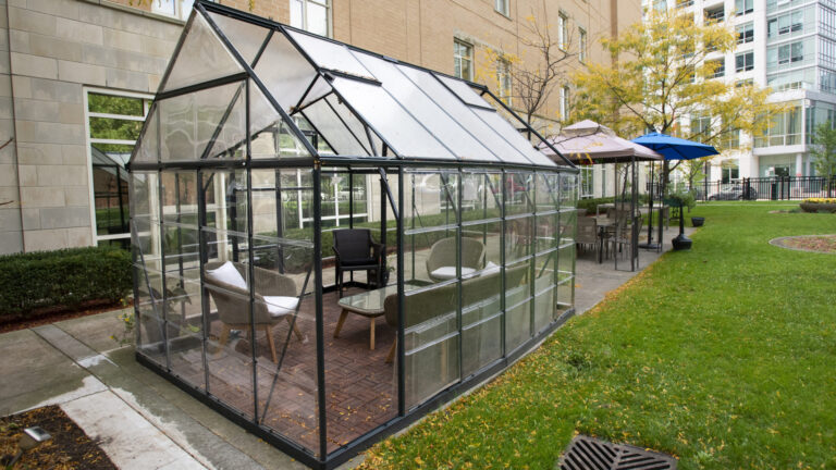 Alongside a four-story brick building, a glass-enclosed structure sits on concrete paving stones. Inside it is a glass coffee table and three chairs, and several of the glass panels are open. Next to it is a small open-air tent with a table and four chairs, along with a third sun umbrella nearby. Next to all three is a grassy area, with a high-rise building at the end of the property.