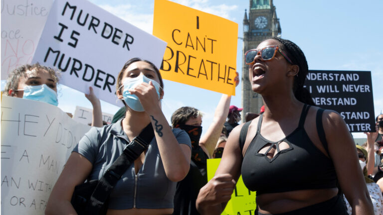 Two young women in front of a series of protest signs saying “I can’t breathe” and “Murder is murder” among others with the Parliament clock tower in the background.
