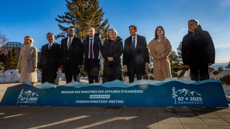 The eight ministers are bundled up against the cold with a sign for their meeting in front of them and a bright blue sky behind them.